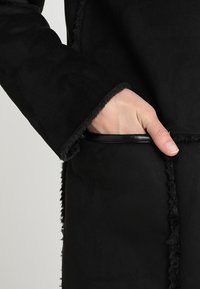 Blazer de ante negro con un acabado texturizado, costuras visibles y un bolsillo con borde de cuero. La tela parece suave y lisa.