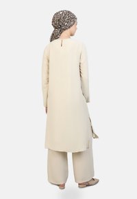 Beige, textured long tunic with side slits, paired with wide-leg pants. Brown patterned scarf tied at the back, flat shoes visible.
