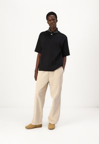 Black oversized t-shirt, beige textured trousers, and tan shoes. Black bandana around the neck, standing against a white background.