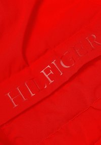 Röd vadderad tyg med en slät textur och ett framträdande präglat "HILFIGER"-logotyp i silver på ena sidan.