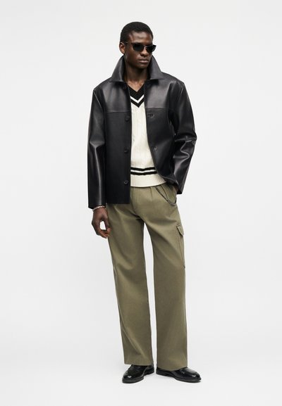 Άνδρας που φοράει μαύρο δερμάτινο μπουφάν, λευκό πουλόβερ με V λαιμόκοψη και μαύρες ρίγες, χακί παντελόνι cargo, μαύρα παπούτσια και γυαλιά ηλίου.