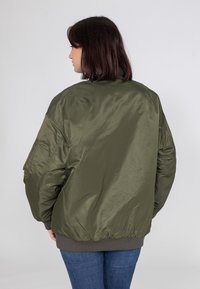 Veste bomber vert olive avec une texture lisse et brillante, poignets et ourlet côtelés, coupe ample, dotée d'une poche latérale sur la manche.