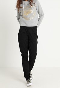 Grijze cropped hoodie met goud logo-ontwerp, gecombineerd met zwarte cargobroeken met zijzakken, en zwart-wit geblokte instapschoenen.