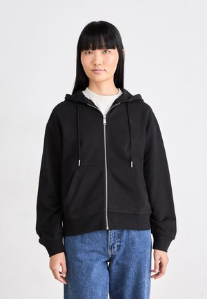 Black zip-up hoodie in fleece material, with a hood, drawstrings, and front pockets. Worn with blue jeans; features a small logo on the chest.