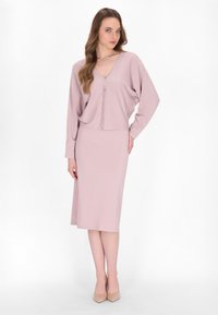 Completo due pezzi rosa composto da una maglietta a maniche lunghe con scollatura a V e bottoni, abbinata a una gonna al ginocchio. Il materiale sembra morbido con una finitura testurizzata.