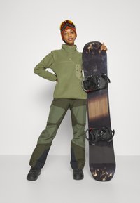 Grüner Fleece-Pullover mit Druckknopfverschluss, kombiniert mit grünen und dunkeloliven Schneehosen. Schwarzes Snowboard mit Bindungen. Orangefarbene Skibrille und Mütze.
