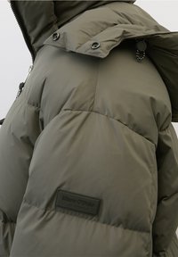 Close-up of olive-green padded winter coat with visible hood, black snaps, zipper, and Marc O'Polo logo patch.