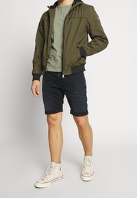 Olivgrön zip-up jacka med huva, kombinerad med en ljusgrön t-shirt och mörka denimshorts. Vita hög-top sneakers fulländar looken.