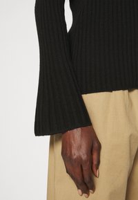 Pull en maille côtelée noire avec manches évasées, associé à un pantalon beige clair. La texture est douce, mettant en valeur des rayures verticales.