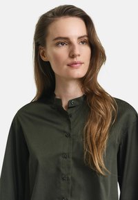 Camicia button-up verde scuro con colletto alla cinese, caratterizzata da una texture liscia e chiusure con bottoni neri lungo la parte anteriore.
