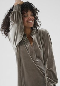 Lächelnde Frau mit lockigem Haar, die ein langärmliges, graues Samtkleid mit gerafften Bündchen und einem Rüschenausschnitt trägt und mit einem erhobenen Arm posiert.