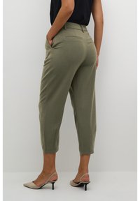 Pantalon vert sur mesure à taille haute, coupe raccourcie, poches avant et texture lisse. Associé à des mules à talons nude, présenté de dos.