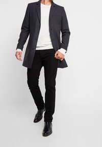 Šedý vlněný kabát na míru se třemi knoflíky, klopovým límcem a náprsní kapsou, nošený přes krémový svetr a černé úzké kalhoty.