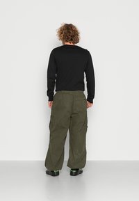 Zwart shirt met lange mouwen en olijfgroene cargobroek met meerdere zakken, losse pasvorm en zwarte schoenen met groene accenten.