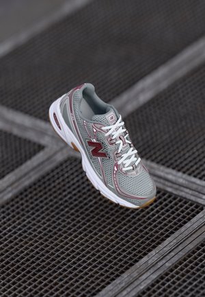 Grå og kastanjefarvet New Balance løbesko med hvide snørebånd, der hviler på en sort metalristet overflade.