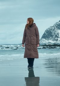 Brauner langer Mantel mit Kapuze, Reißverschluss und zwei vorderen Taschen, getragen über schwarzen Hosen, an einem Strand mit felsiger Küste und bewölktem Himmel.