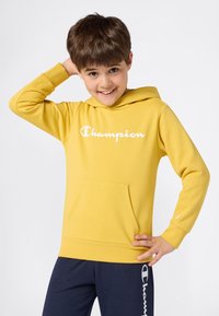 Młody chłopiec uśmiechający się z ręką za głową, ubrany w żółtą bluzę Champion i granatowe dresy z białymi logo Champion.