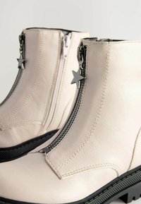 Stivali alla caviglia in pelle bianca con doppie cerniere e accenti a forma di stella, superficie strutturata, suola in gomma nera e dettagli di cucitura in contrasto.