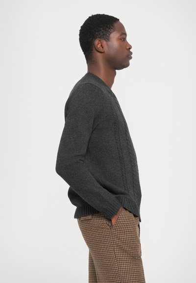 Pull en tricot gris foncé avec un motif en cablage, présentant un col rond et des poignets côtelés, associé à un pantalon en pied-de-poule marron.
