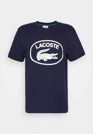 Granatowy bawełniany T-shirt z białym logo krokodyla i napisem „LACOSTE” w owalnym wzorze. Krótkie rękawy i okrągły dekolt.