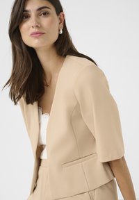 Beige bleiseri, joka on räätälöity lyhyillä hihoilla ja avoimella edessä, jossa on sileä pinta ja hienovarainen kiilto. Sisältää sivutaskut ja siistit linjat.