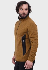 Braune Fleecejacke mit strukturierter Oberfläche, ausgestattet mit einer schwarzen Seitentasche mit Reißverschluss und einem hohen Kragen. Angesetzter Reißverschluss und kontrastierende Manschetten.