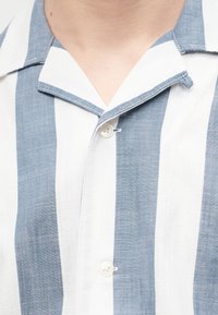 Nærbillede af blå og hvide vertikale stribede skjorte med krave, knappet ved brystet.