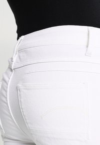 Valkoiset farkut, joissa on tiukka muotoilu, takatasku ja pieni tekstuuri. Saumojen väritys on vaalea ja näkyvä.