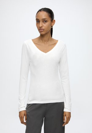 Femme aux cheveux raides et foncés portant un haut blanc ajusté à manches longues avec col en V et un pantalon gris, debout devant un fond blanc uni.