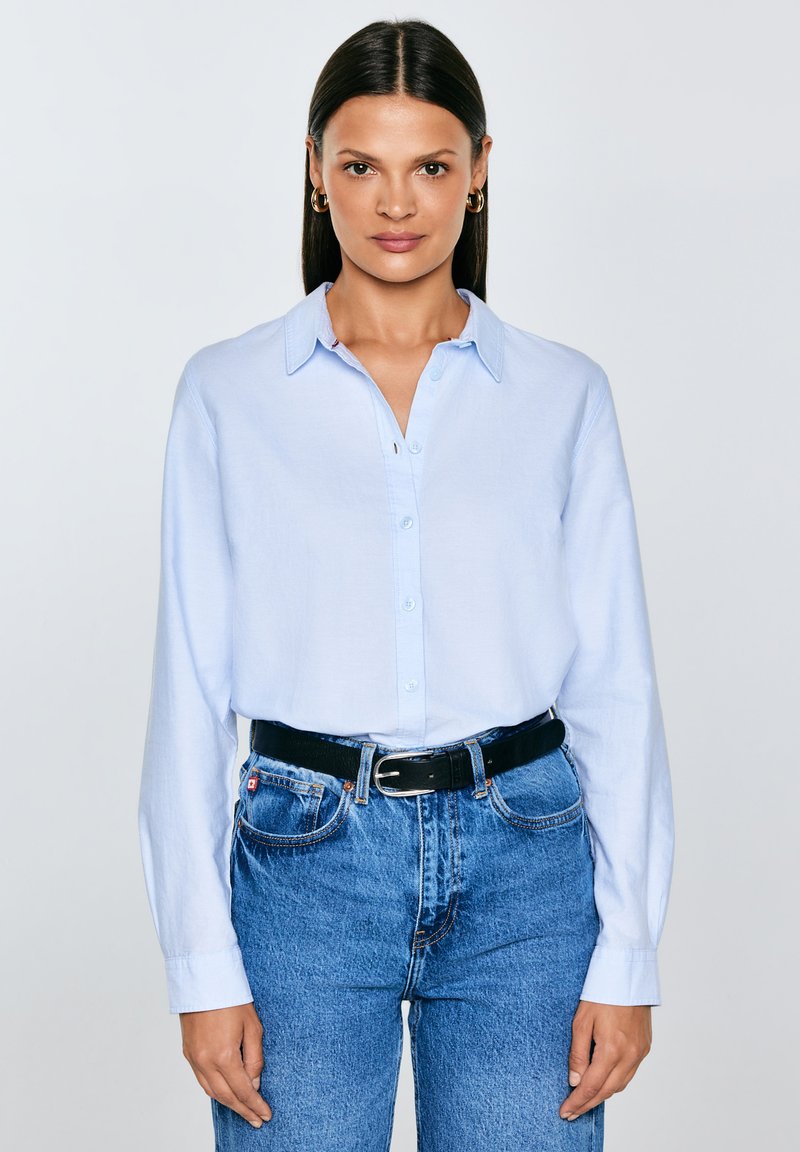 Sieviete ar taisniem tumšiem matiem valkā zelta riņķa auskarus, gaiši zilu kreklu ar pogām, tumšu jostu un augstvēderu zilu džinsu bikses, skatās uz priekšu.