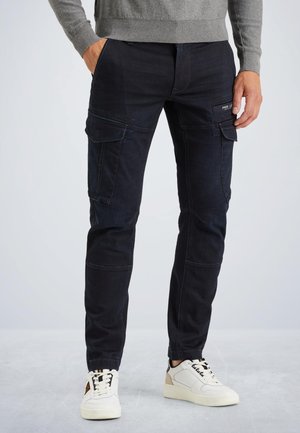 Mann trägt schwarze Cargo-Hosen mit Seitentaschen, grauen Pullover und weiße Sneakers, steht auf einem hellgrauen Boden vor einem schlichten Hintergrund.