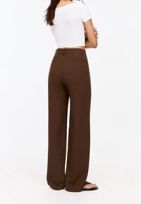 Pantalon large marron taille haute, coupe ajustée et tissu lisse. Associé à un crop top blanc moulant et des tongs noires.
