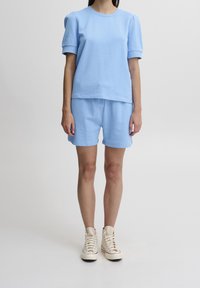 Kvinna som bär en ljusblå kortärmad topp och matchande shorts, stående, i kombination med krämfärgade hög-top sneakers mot vit bakgrund.