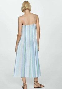 Donna con capelli biondi corti che indossa un vestito midi a strisce senza maniche nei colori blu, verde e bianco, abbinato a sandali piatti con cinturini marroni, vista di spalle.