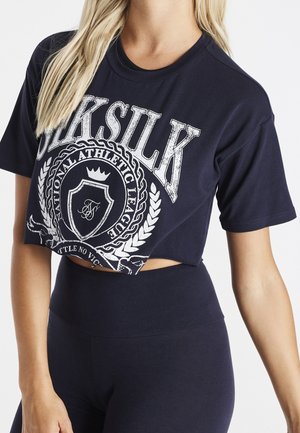Kobieta w granatowym krótkim T-shircie z białym logo "SikSilk" i sportowym herbem, w połączeniu z granatowymi legginsami z wysokim stanem.