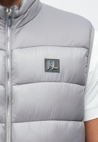 Siva brezrokavka s šivi in sprednjim zadrgo ter črnim kvadratnim logotipom na prsih, nošena preko belega kratkoročnega majice.