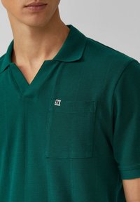 Dunkelgrünes Poloshirt aus strukturiertem Material, mit einem umgeschlagenen Kragen und einer Brusttasche mit kleinem Logo.