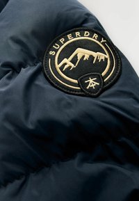 Jūras zilas pūkainas jakas sadaļa ar apaļu, izbrodētu melnu un zeltītu uzplecu ar kalnu dizainu un tekstu "SUPERDRY".