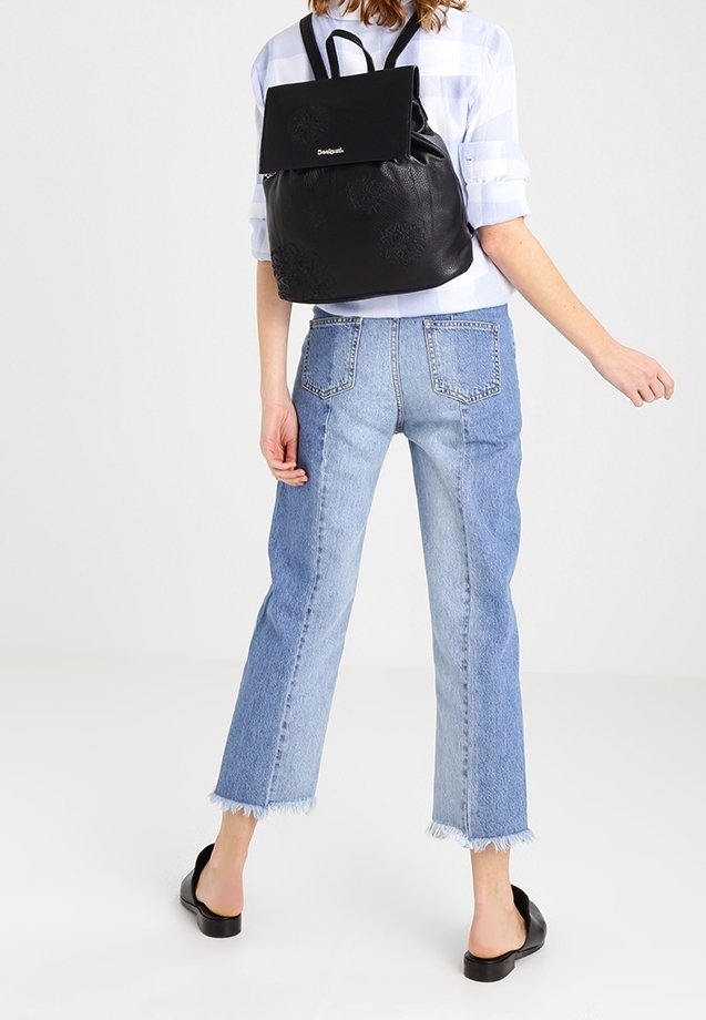 Schwarzer Leder-Rucksack mit Klappe und Logo, kombiniert mit hellblauen, kürzeren Jeans mit ausgefransten Säumen und schwarzen flachen Schuhen.