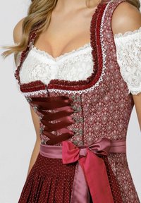 Burgundy dirndl with white lace top, intricate floral pattern, satin lace-up front, and a bow at the waist. Pleated skirt with polka dots.