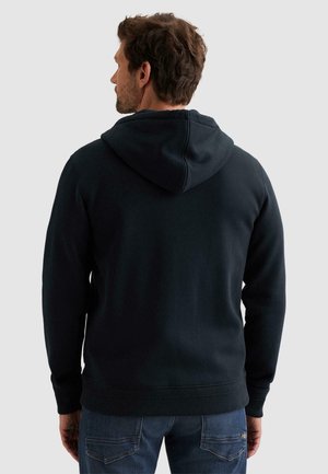 Homme portant un sweat à capuche noir uni et un jean bleu, vu de dos sur un fond gris clair.