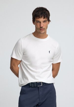 T-shirt bianca in cotone con collo rotondo, caratterizzata da un piccolo logo blu navy sul petto sinistro, abbinata a pantaloni blu navy e una cintura intrecciata.