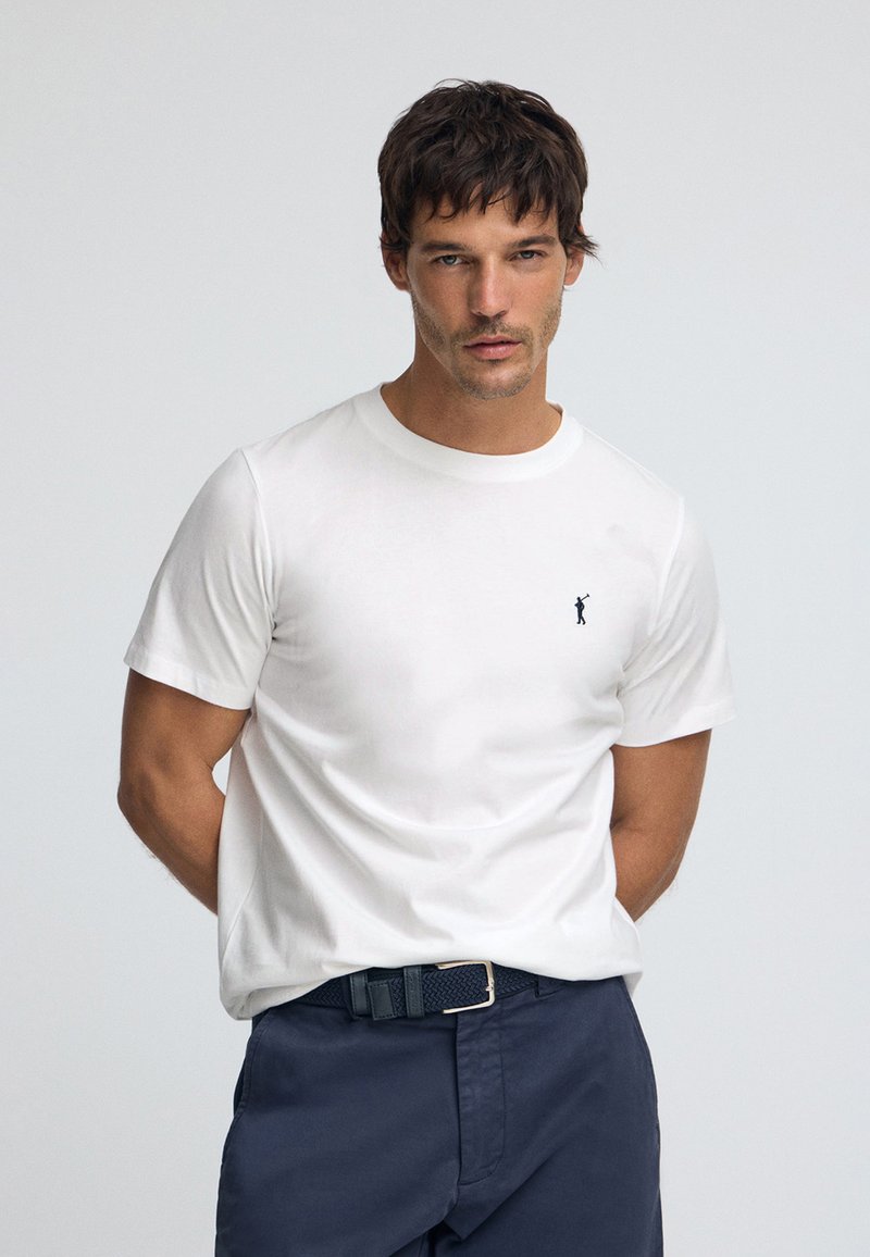 Biały bawełniany T-shirt z okrągłym dekoltem, z małym granatowym logo na lewej piersi, zestawiony z granatowymi spodniami i plecionym paskiem.