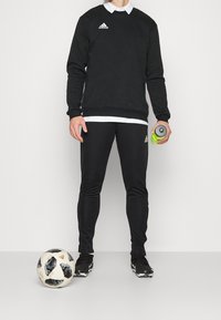 Sudadera negra con cuello blanco, pantalones negros y zapatillas deportivas negras. Sosteniendo una botella de agua verde junto a un balón de fútbol.