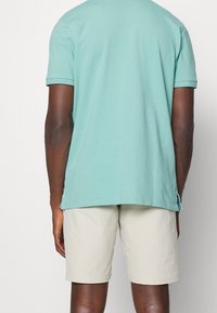 Světle modrá bavlněná trička s krátkým rukávem, s zaobleným lemem a bočními rozparky, spárované s béžovými šortkami, z pohledu zezadu.