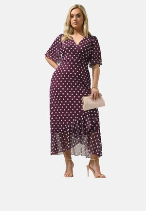 Kvinde iført lilla polkaprik-wrapkjole, der holder en beige clutch, kombineret med nøgenfarvede hæle, stående mod en hvid baggrund.
