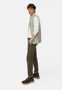 Hellgraue Weste mit Armausschnitten, schwarzer Reißverschluss, weißes Hemd, olivgrüne Hose, braune Wanderstiefel, stehende Pose mit der linken Hand in der Tasche.