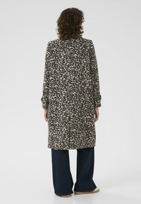 Une personne aux cheveux bouclés porte un long manteau en imprimé léopard, un pantalon large sombre et des chaussures claires, se tenant de dos sur un fond blanc.