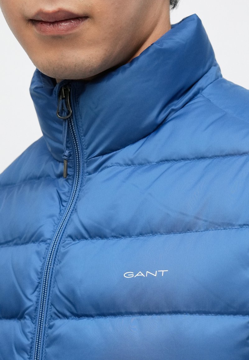 Blue puffer jacket with a high collar. Features quilted texture, a front zip, and a white logo on the left side. Smooth, lightweight fabric.
