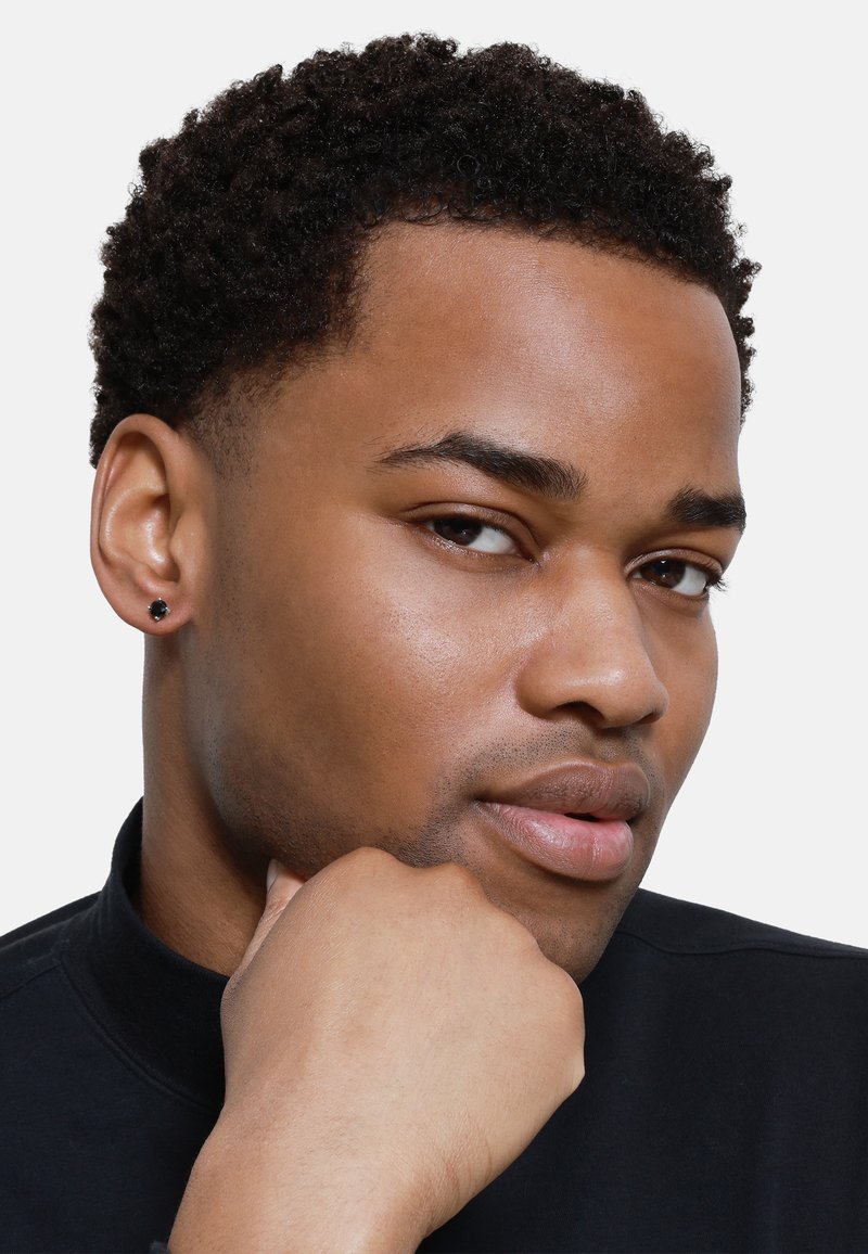 Boucles d'oreilles noires sur une personne ayant les cheveux courts et bouclés, portant un haut noir à col montant. L'arrière-plan est blanc uni, mettant en valeur les traits du visage.